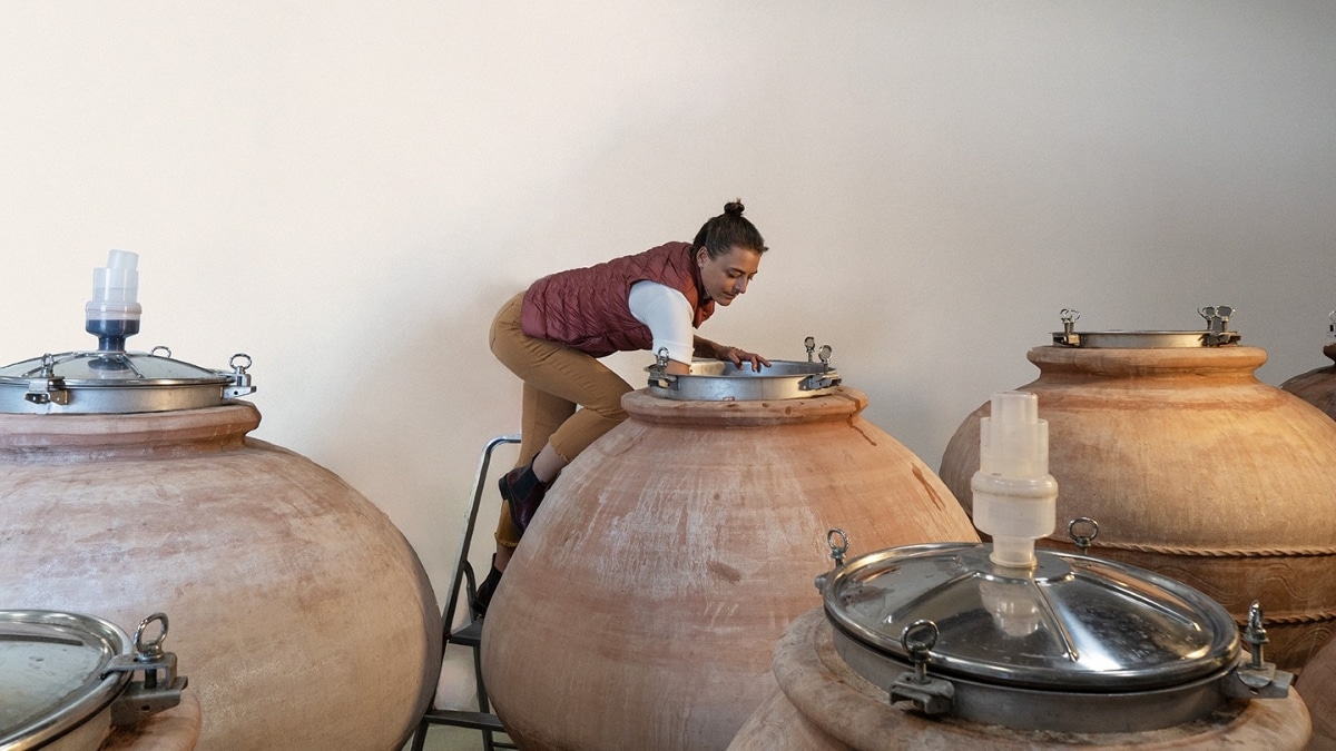 Elena Casadei inspecting a qvevri in a cellar, surrounded by large clay vessels used for wine fermentation and ageing.