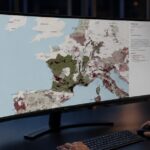 Man at a desk viewing Open Wine Map on a curved monitor, showing European wine regions in shades of green and burgundy, with filter options for appellations.